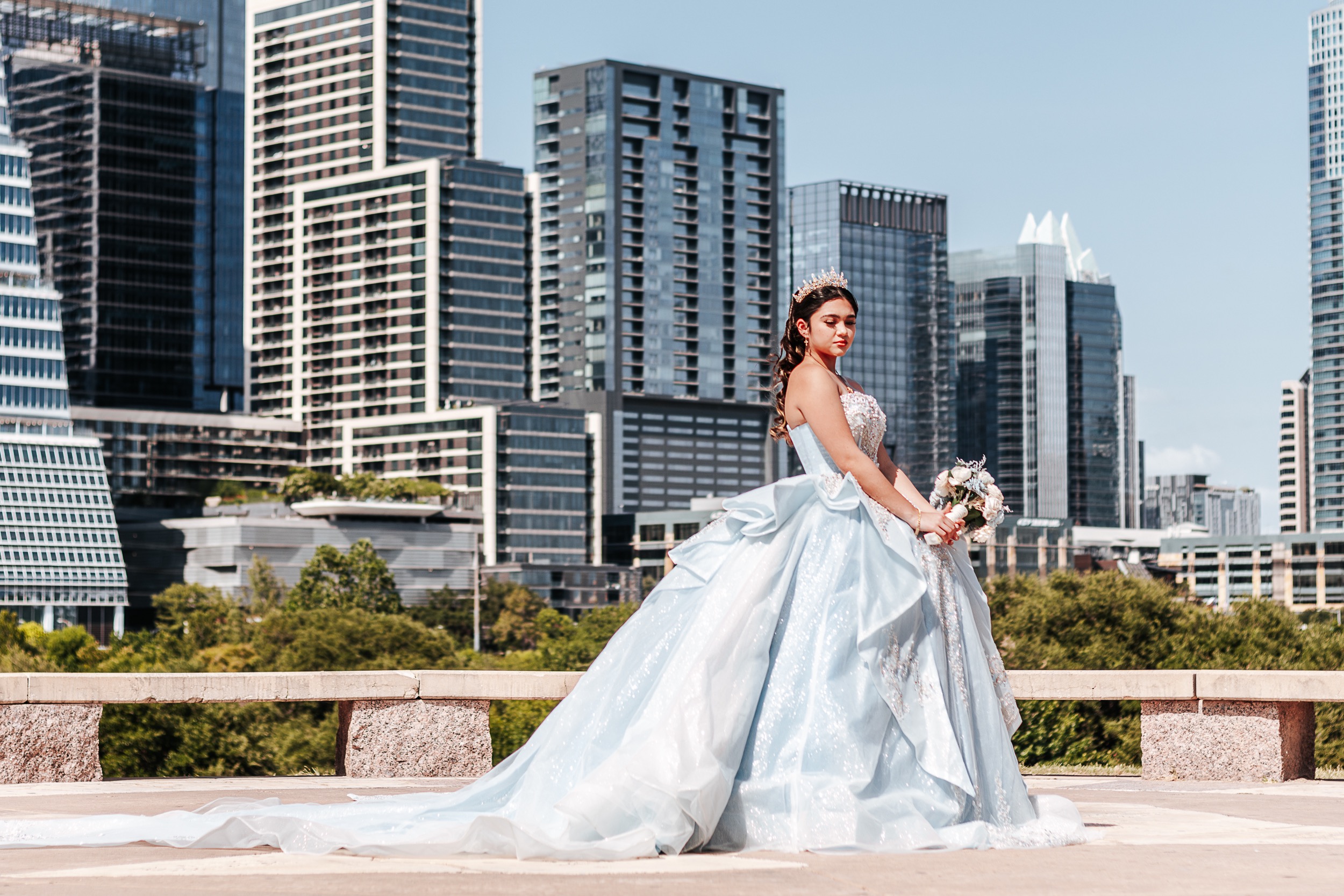Quinceañera in a light blue ball gown holding a bouquet with the Austin skyline behind her, photographed by Weddings by Wendi