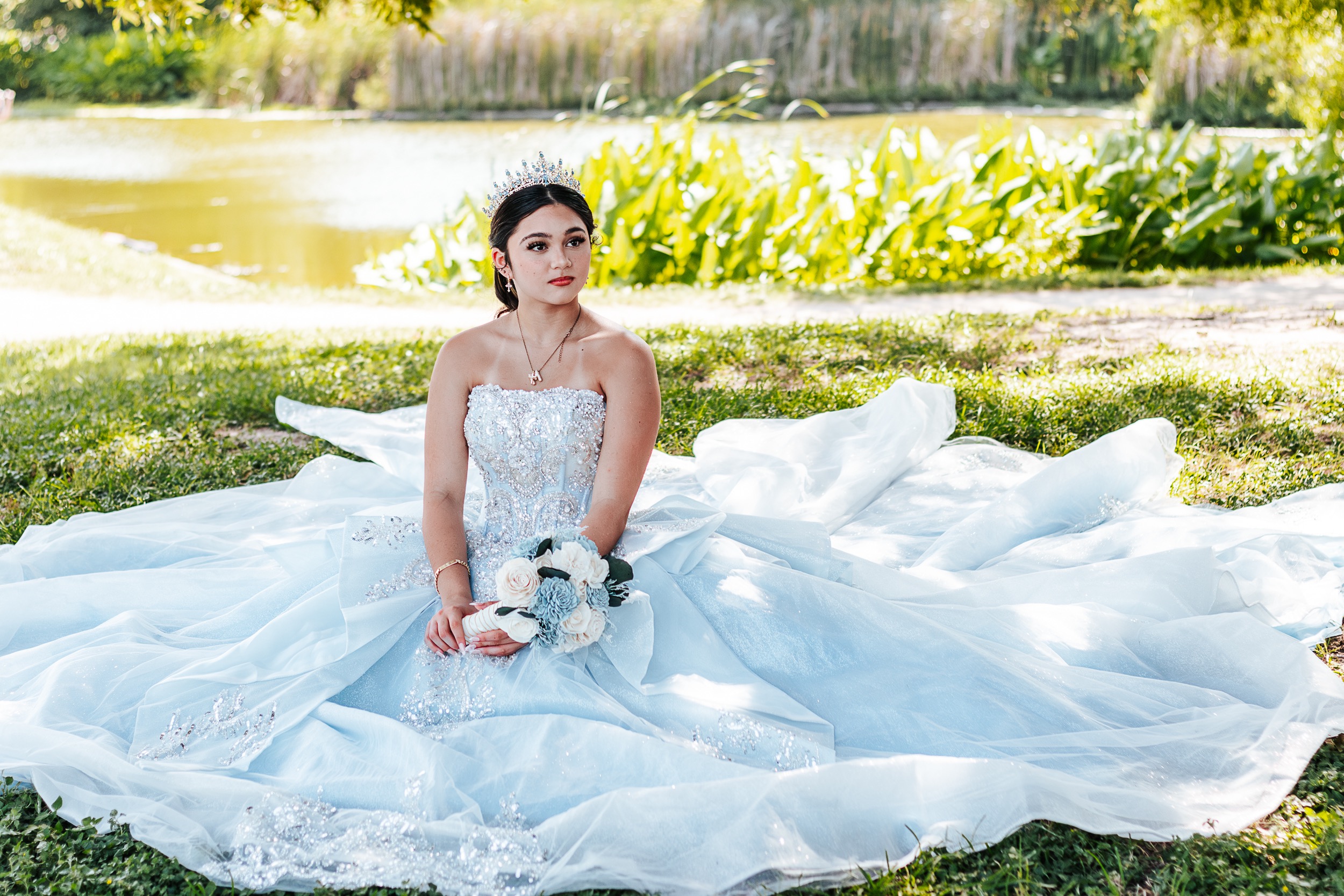 Quinceañera seated on the grass beside a pond in a light blue gown with her dress spread around her, photographed by Weddings by Wendi