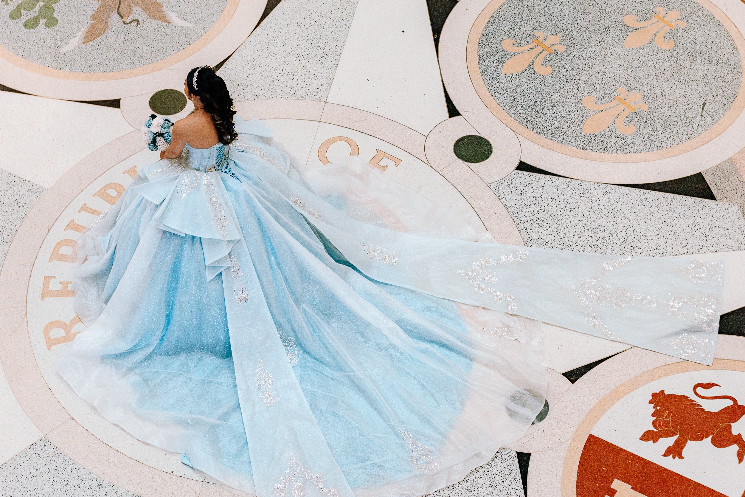 Aerial view of a quinceañera's light blue gown spread across the medallion floor of the Texas State Capitol rotunda, photographed by Weddings by Wendi
