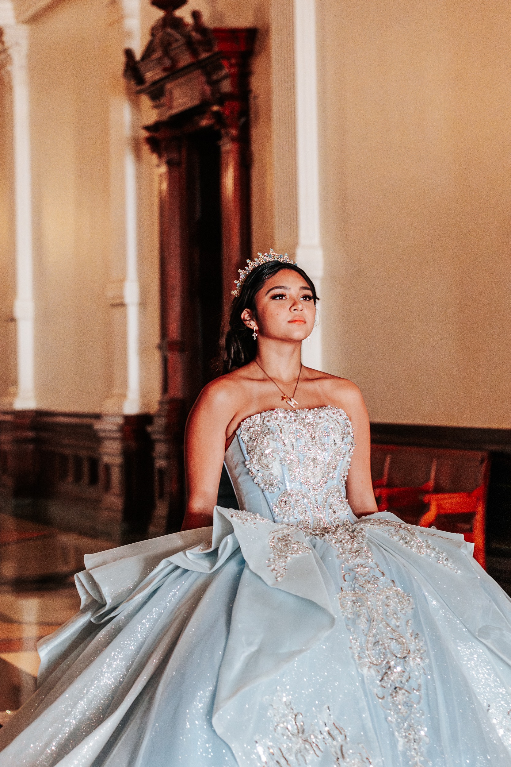 Quinceañera in a beaded light blue gown and tiara inside the Texas State Capitol between marble columns, photographed by Weddings by Wendi