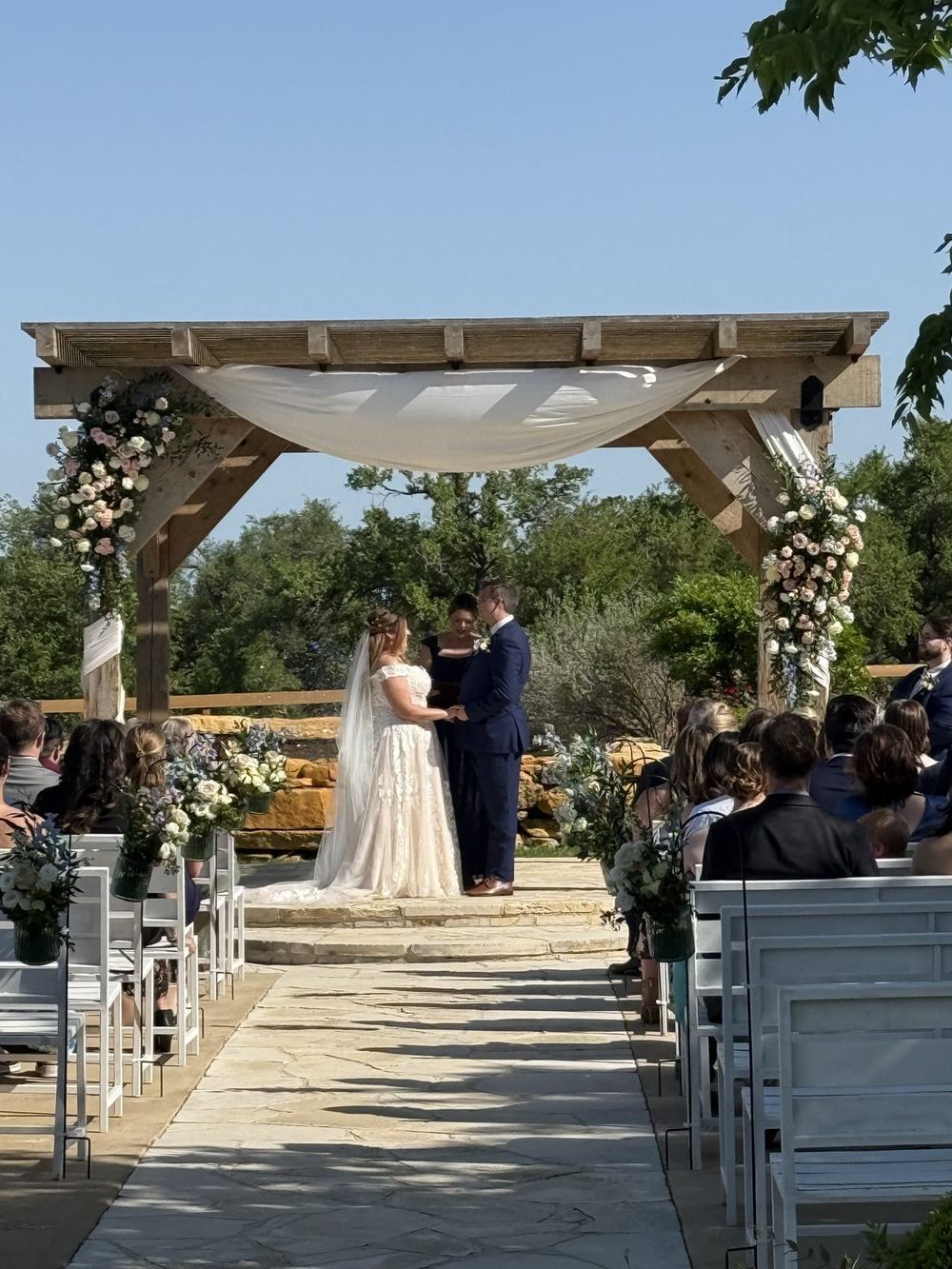 Bride and groom exchanging vows beneath a floral-draped wooden pergola at Memory Lane Ranch in Dripping Springs, Texas